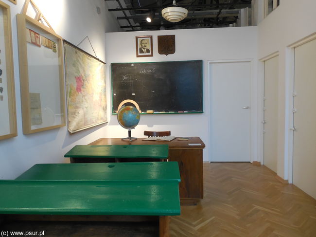 Szkolna sala: zielone ławki, tablica i portret Bieruta